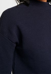 Granatowy sweter z prążkowanego dzianiny o wysokim dekolcie i teksturowanej tkaninie. Posiada opuszczone szwy ramion, co zapewnia luźny krój.