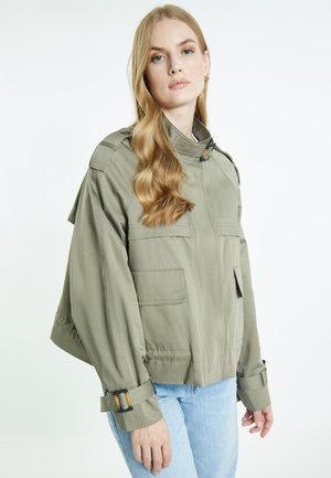 Donna con lunghi capelli biondi che indossa una giacca utilitaria verde chiaro e jeans azzurri su sfondo bianco semplice.