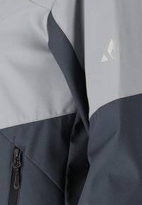 Gros plan sur la manche d'une veste gris foncé et gris clair avec une fermeture éclair et un petit logo triangulaire sur le haut du bras.