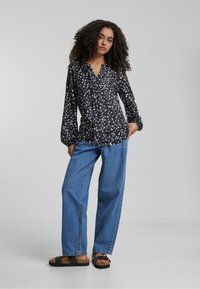 Svart bluse med blomsterprint, V-hals og lange ermer, parret med løse blå jeans og svarte platåsandaler på en grå bakgrunn.