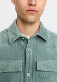 Groene corduroy overhemd met een omgeslagen kraag, twee borstzakken en knoopsluiting. De stof heeft een textuur met zichtbare ribbels.