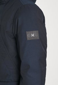 Marinblå puffer-jacka med strukturväv, med en framträdande logopatch på vänster ärm i svart och silverdetaljer.