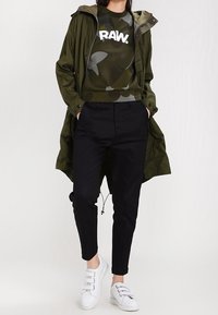 Veste légère vert olive portée sur un sweat-shirt camouflage sombre avec le texte "RAW." Pantalons noirs fuselés et baskets blanches complètent la tenue.