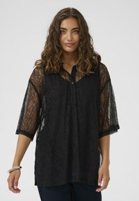 Femme aux cheveux bruns bouclés portant un blouse en dentelle noire à manches trois-quarts et un pantalon sombre, se tenant devant un fond uni.