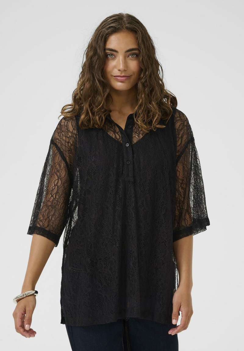 Femme aux cheveux bruns bouclés portant un blouse en dentelle noire à manches trois-quarts et un pantalon sombre, se tenant devant un fond uni.