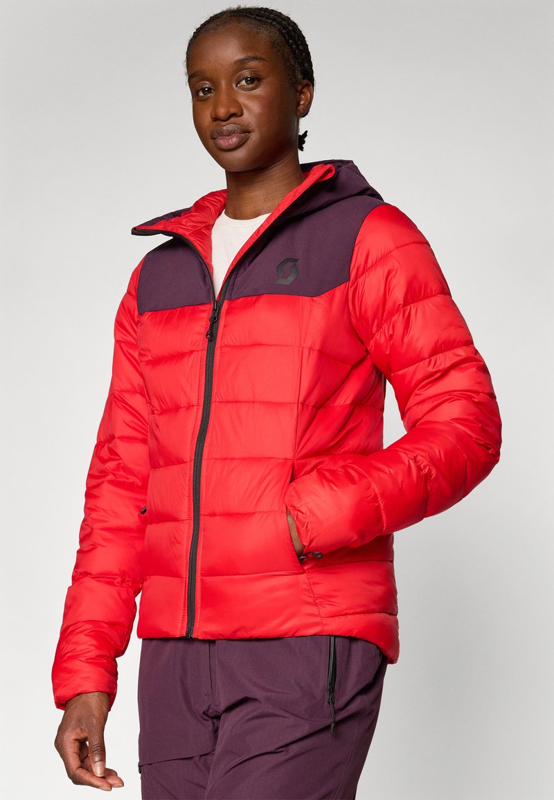 Czarna kobieta w czerwonej i ciemnofioletowej kurtce ocieplanej z kapturem oraz fioletowych spodniach, stojąca na jednolitym jasnoszarym tle.