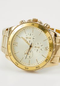 Montre dorée avec un boîtier rond, un cadran lisse texturé avec trois sous-cadrans et un bracelet maillon. Les indications incluent des chiffres et des marqueurs de minutes.