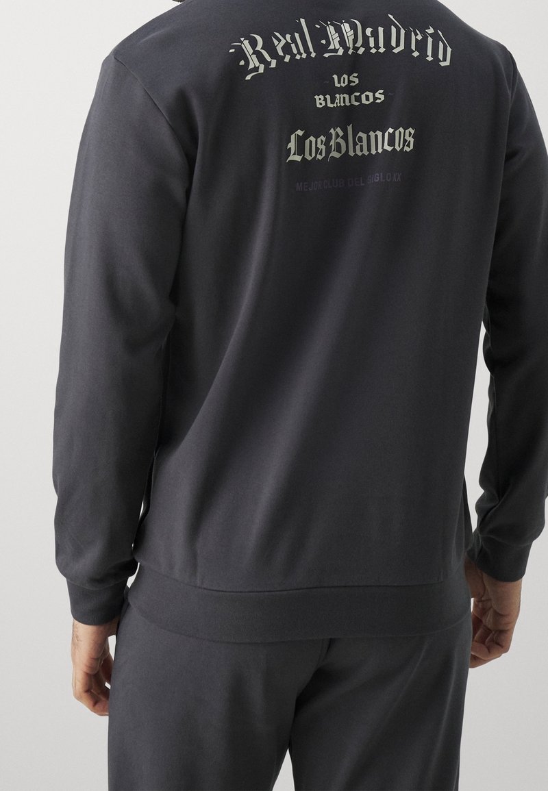 Felpa grigia scura con maniche lunghe, con la scritta "Real Madrid" e disegni sul retro, realizzata in tessuto morbido con una vestibilità comoda.