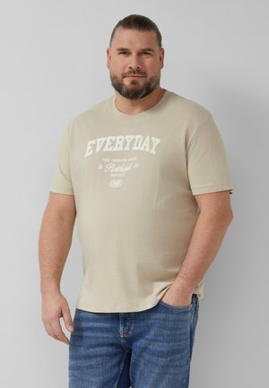 Mężczyzna z brodą, w beżowej koszulce z napisem "EVERYDAY" i niebieskich dżinsach, stoi na tle jednolitego jasnoszarego tła z jedną ręką w kieszeni.