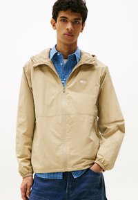 Beżowa kurtka z kapturem o gładkiej fakturze, z przodu zapinana na zamek, z elastycznymi mankietami, noszona na niebieskiej koszuli w kratę, z małą naszywką logo.