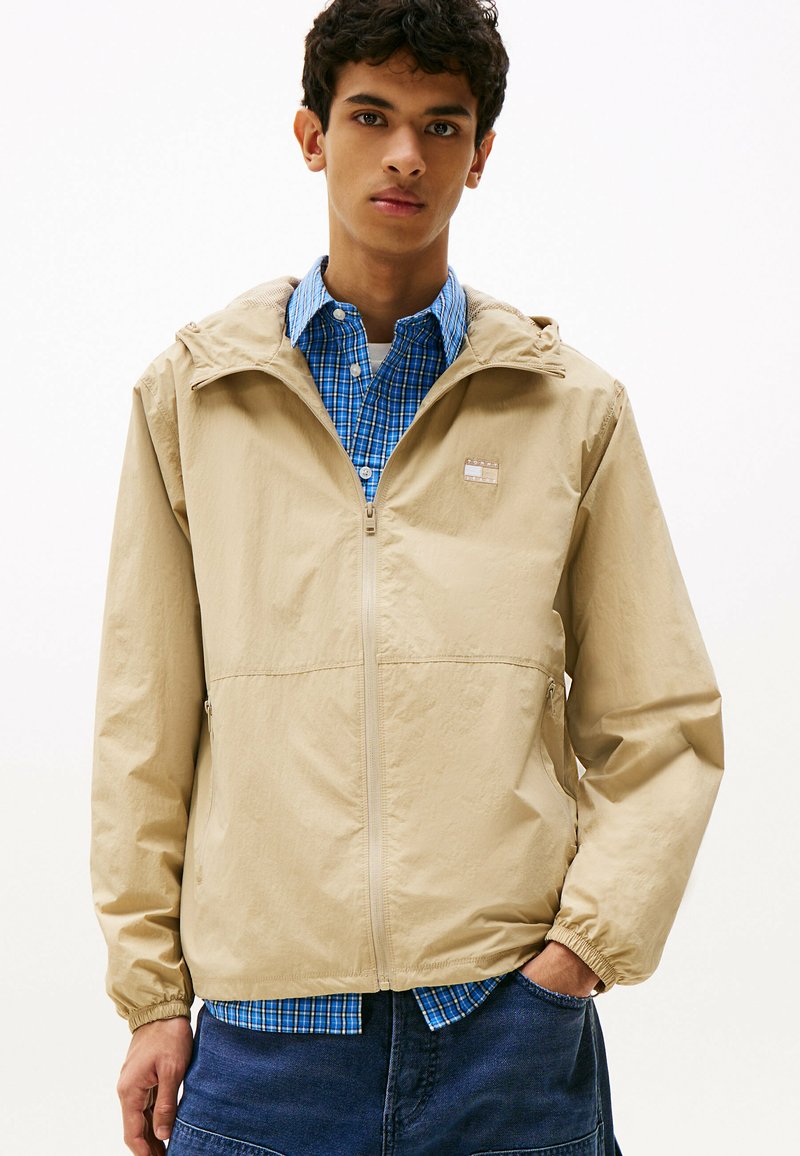 Beżowa kurtka z kapturem o gładkiej fakturze, z przodu zapinana na zamek, z elastycznymi mankietami, noszona na niebieskiej koszuli w kratę, z małą naszywką logo.