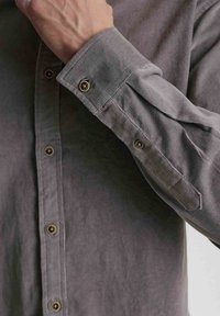 Grå skjorte med knaplukning og en glat tekstur, der har guldtonede knapper, en enkelt manchet og et strømlinet design.