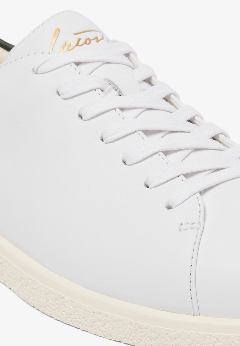 Primo piano di una sneaker in pelle bianca con lacci bianchi e suola in gomma testurizzata color avorio, con scritta in oro in caratteri corsivi vicino alla linguetta.