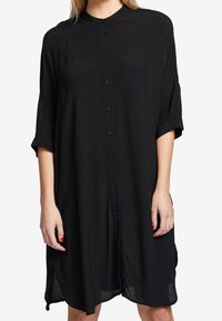 Svart knappuppklädd klänning med krage, korta ärmar och sidokläppar. Tillverkad av lätt material med en mjuk textur. Längden når mitt på låret.