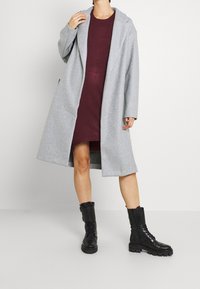 Donna che indossa un cappotto grigio fino al ginocchio sopra un vestito bordeaux con stivali militari neri con lacci, in piedi di fronte a uno sfondo semplice.