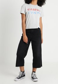 Camiseta gris recortada con el logo "TOMMY" en rojo y azul, combinada con jeans negros de pierna ancha y zapatillas negras de caña alta.
