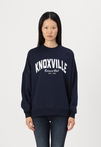 Tamsiai mėlynas džemperis su baltu užrašu "KNOXVILLE Leisure Club EST. 1982", atsipalaidavęs siluetas, briaunoti apvadai ir nuleistos pečių siūlės.
