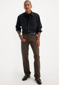 Camisa negra de manga larga con dos bolsillos en el pecho, combinada con pantalones marrones y zapatos negros. El conjunto presenta un diseño clásico y relajado.