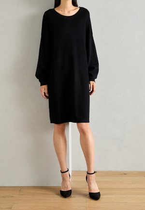 Vrouw draagt een loszittende, lange mouwen zwarte jurk met een ronde hals en zwarte enkellaarsjes met riempje, staand op een houten vloer.