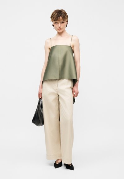 Νεαρό άτομο φοράει φαρδιά ελιάς πράσινη μπλούζα χωρίς μανίκια, φαρδιά μπεζ παντελόνια, μαύρα παπούτσια με μυτερή μύτη, κρατώντας μαύρη τσάντα, στέκεται μπροστά σε απλό φόντο.