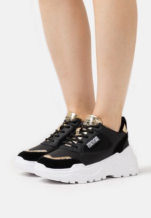 Schwarze und goldene Sportsneaker aus Mesh und Wildleder, mit einer klobigen weißen Sohle und auffälligem Branding auf der Zunge.