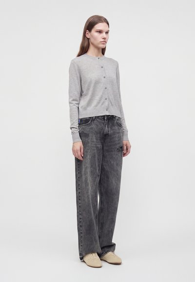 Jeune personne debout portant un cardigan gris clair boutonné, un jean noir délavé et ample, et des chaussures beige sur un fond clair uni.