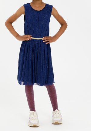 Robe sans manches marine avec des accents scintillants, jupe froncée et ceinture dorée. Assortie à des leggings scintillants violets et des baskets blanches.