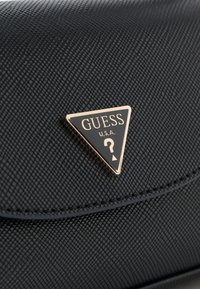 Didelis priartinimas prie tekstūruotos juodos medžiagos su auksiniu ir juodu trikampiu Guess U.S.A. logotipo plokštelė ant rankinės arba maišo.