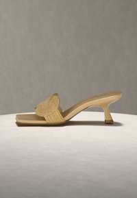 Bézs színű, nyitott orrú papucs közepes magasságú, ívelt sarokkal és széles, kötött pánttal, mely csomó részlettel díszített, semleges felületen és háttér előtt elhelyezve.