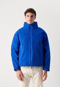 Giacca impermeabile blu con colletto alto, chiusura a zip, due tasche laterali con zip e polsini elasticizzati. Materiale morbido e leggero.