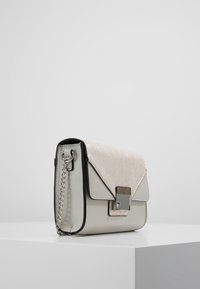 Metalická stříbrná kabelka s texturovaným horním klopou, řetízkovým popruhem, čtvercovým uzávěrem a hladkým spodním materiálem. Kompaktní obdélníkový tvar.