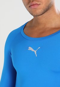 Maglietta atletica a maniche lunghe blu realizzata in materiale elastico, con scollatura rotonda e logo riflettente argento sul petto.