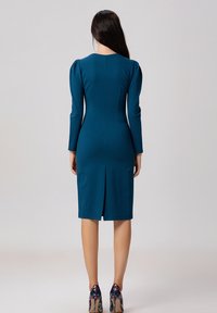 Robe ajustée teal avec manches longues, décolleté rond et fente à l'arrière. Texture lisse, design structuré et coupe cintrée. Chaussures colorées visibles.