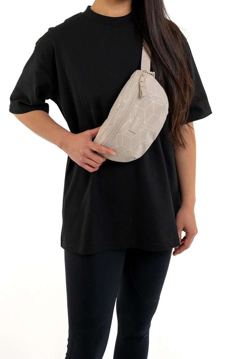 Personne portant un t-shirt noir ample et un pantalon noir, tenant un sac bandoulière beige avec des motifs géométriques sur la poitrine.