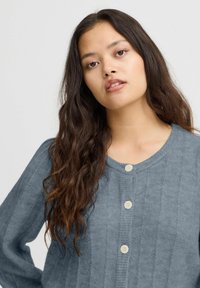 Šviesiai mėlynas mezginys kardiganas su įlinkta tekstūra, apvalia iškirpte ir penkiomis grietinėlės spalvos sagomis priekyje. Ilgos rankovės su laisva prigludimo forma.