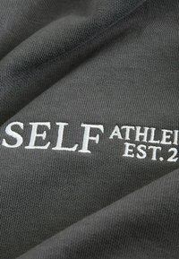 Szary materiał o prążkowanej fakturze, z białym haftowanym napisem "SELF ATHLETE EST. 2."