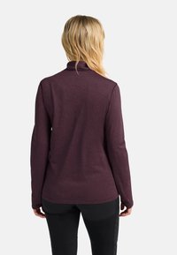 Donna bionda che indossa una giacca lunga a maniche, di colore viola scuro, e pantaloni neri, vista da dietro su uno sfondo bianco uniforme.