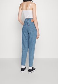 Högmidjade blå denimjeans med avslappnad passform, försedd med en läderlogotyp, två bakfickor och uppvikta anklar. Svarta sneakers är synliga.