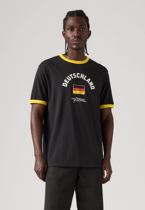 Bărbat cu părul împletit, purtând un tricou negru cu margine galbenă, steagul Germaniei și textul „Deutschland”, stând în fața unui fundal simplu și luminos.