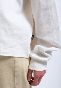 Weißes Langarmshirt aus Baumwolle mit weicher Textur. Verfügt über eine entspannte Passform und einem kleinen Logotag an der Seite. Kombiniert mit beigen Hosen.