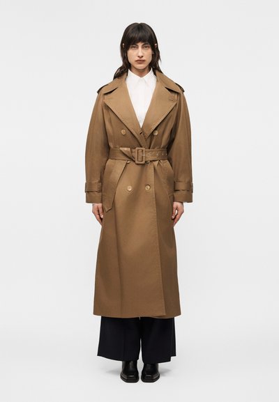 Femme debout, portant un long trench-coat marron ceinturé sur une chemise blanche, un pantalon noir à jambes larges et des chaussures noires à bout carré, sur un fond uni.
