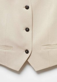 Veste beige sur mesure avec trois boutons foncés au centre et deux poches passepoilées horizontales près de l'ourlet.
