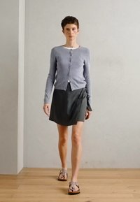 Világosszürke, bordázott kardigán gombokkal, sötét, bugyborékoló szoknyával és barna szandálokkal párosítva. Simító szövet, minimalista dizájn.