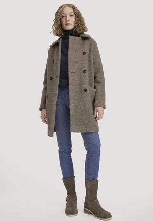 Junge Frau mit lockigem Haar, die einen braunen Wollmantel, einen schwarzen Rollkragenpullover, blaue Jeans und braune Wildlederstiefel trägt, steht vor einem schlichten Hintergrund.