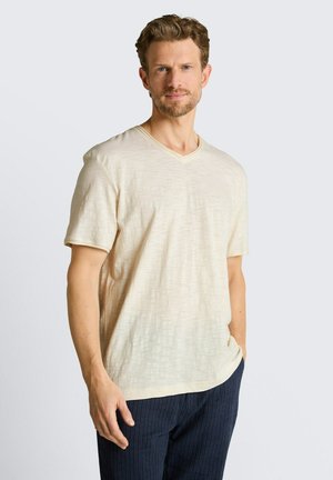 Man met kort bruin haar en baard, die een beige T-shirt met korte mouwen en V-hals en een donkere broek met fijne strepen draagt, staat met één hand in de zak.