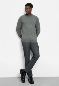 Grauer Strickpullover mit rundem Halsausschnitt, kombiniert mit dunkelgrauer, maßgeschneiderter Hose. Schwarze Lederschuhe vervollständigen das Outfit.