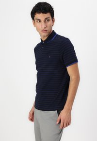 Námořnické polo tričko s tenkými modrými pruhy, krátkými rukávy a dvouknoflíkovou légou. Má malé logo na hrudi a kontrastní límec.