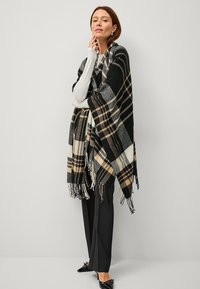 Black and white plaid shawl with beige accents, fringed edges, and a soft texture, draped over a person in black trousers.