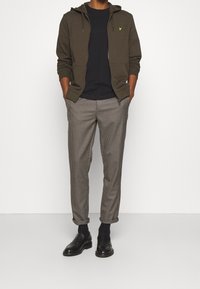 Felpa verde con zip sopra una maglietta blu navy, abbinata a pantaloni grigi a quadri e scarpe di pelle nere; presenta un piccolo logo sulla felpa.