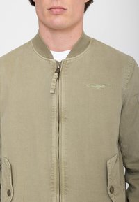 Man draagt een beige bomberjack met rits, geribde kraag, klepzakken met knopen en een klein geborduurd vleugelembleem op de linkerborst.